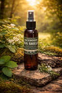 A bottle of BACKWOODS BUG SHIELD Natural Repellent spray stands on a weathered log outdoors, surrounded by moss, green leaves, small white flowers, and soft sunlight filtering through trees.