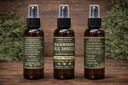 Three amber glass spray bottles of Leaning Oak Soaps botanical outdoor products standing on a wooden surface with a blurred green foliage background. The center bottle is labeled 'Backwoods Bug Shield' and the side bottles show 'Ingredients' and 'How to Use' labels.