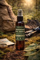 A bottle of Backwoods Bug Shield botanical outdoor spray by Leaning Oak Soaps stands on forest ground with moss, leaves, a backpack, and outdoor gear in a sunlit woodland setting.